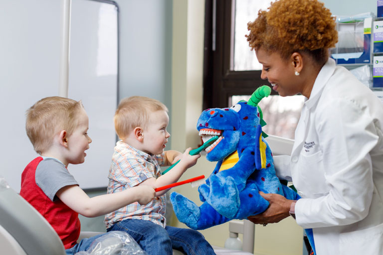 A dentistry professional holds a stuffed dragon as two young children brush its teeth.
