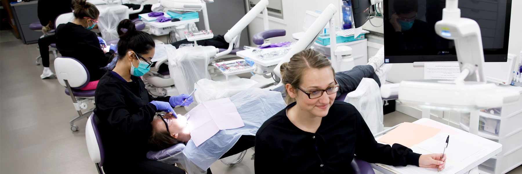 A row of dental hygienists work on patients' teeth as they sit in exam chairs.