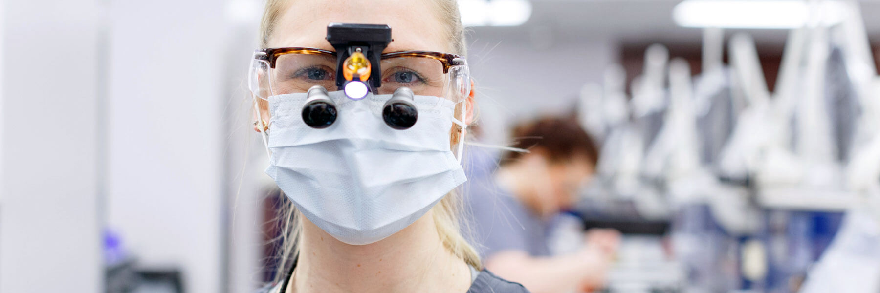 A student wears her scrubs, mask, and protective glasses with headlight.