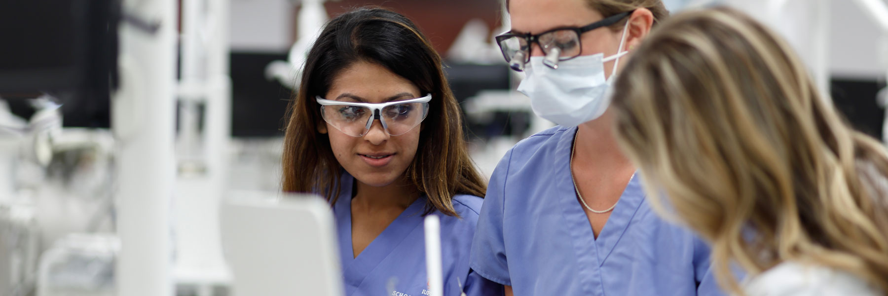 Three students wear scrubs and protective glasses and work together in a lab.