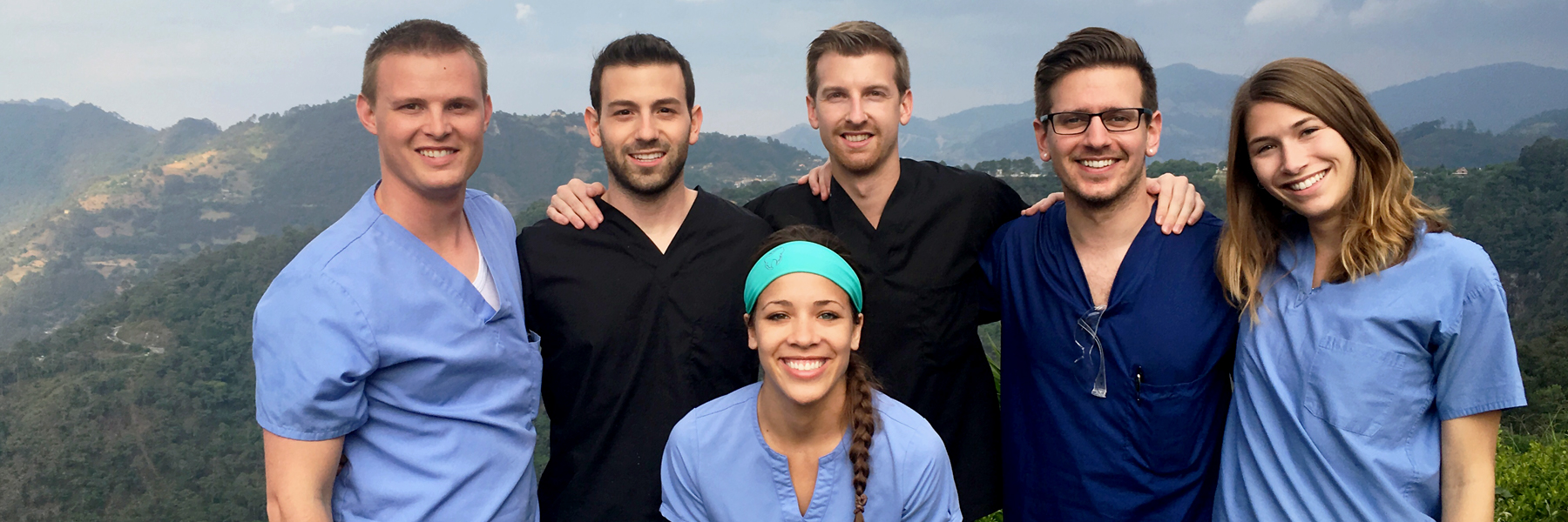 A group of students in their scrubs pose in front of a green, hilly landscape.