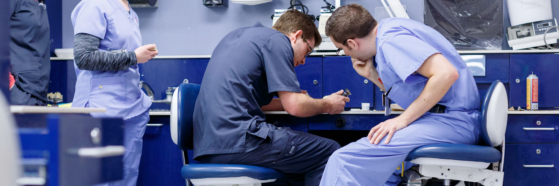 Two male dentistry students in scrubs work with dental tools at a research lab station.