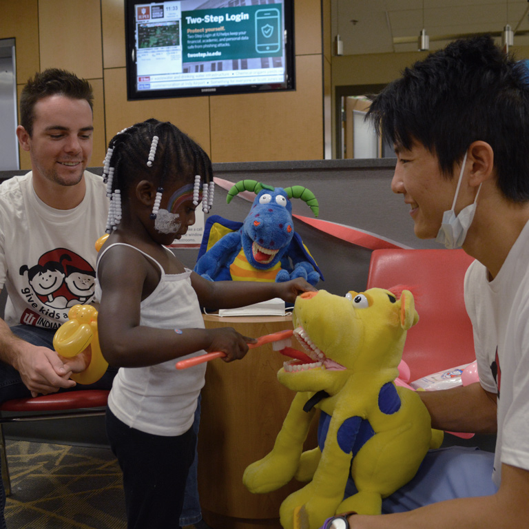 Two dentistry students show a young girl stuffed animals as she practices brushing their teeth.